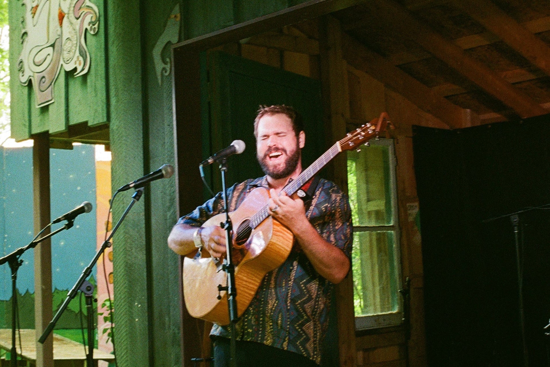 Man playing guitar in front of a wooden cabin with microphones set up.