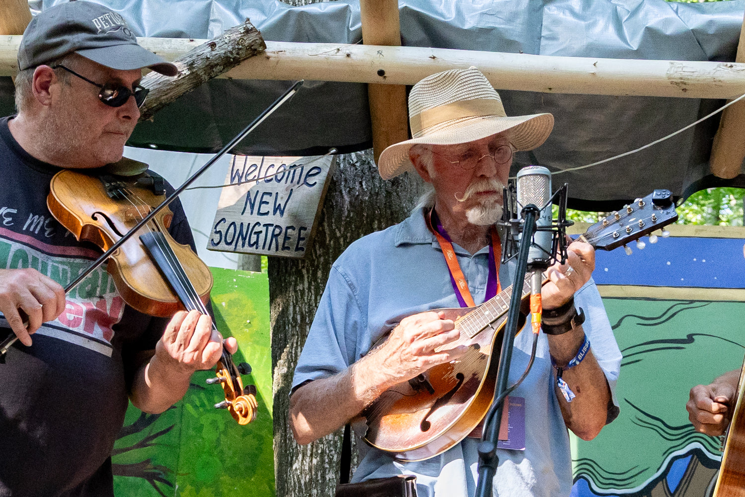 Two musicians playing instruments with a microphone and 'Welcome New Songtree' sign in the background.