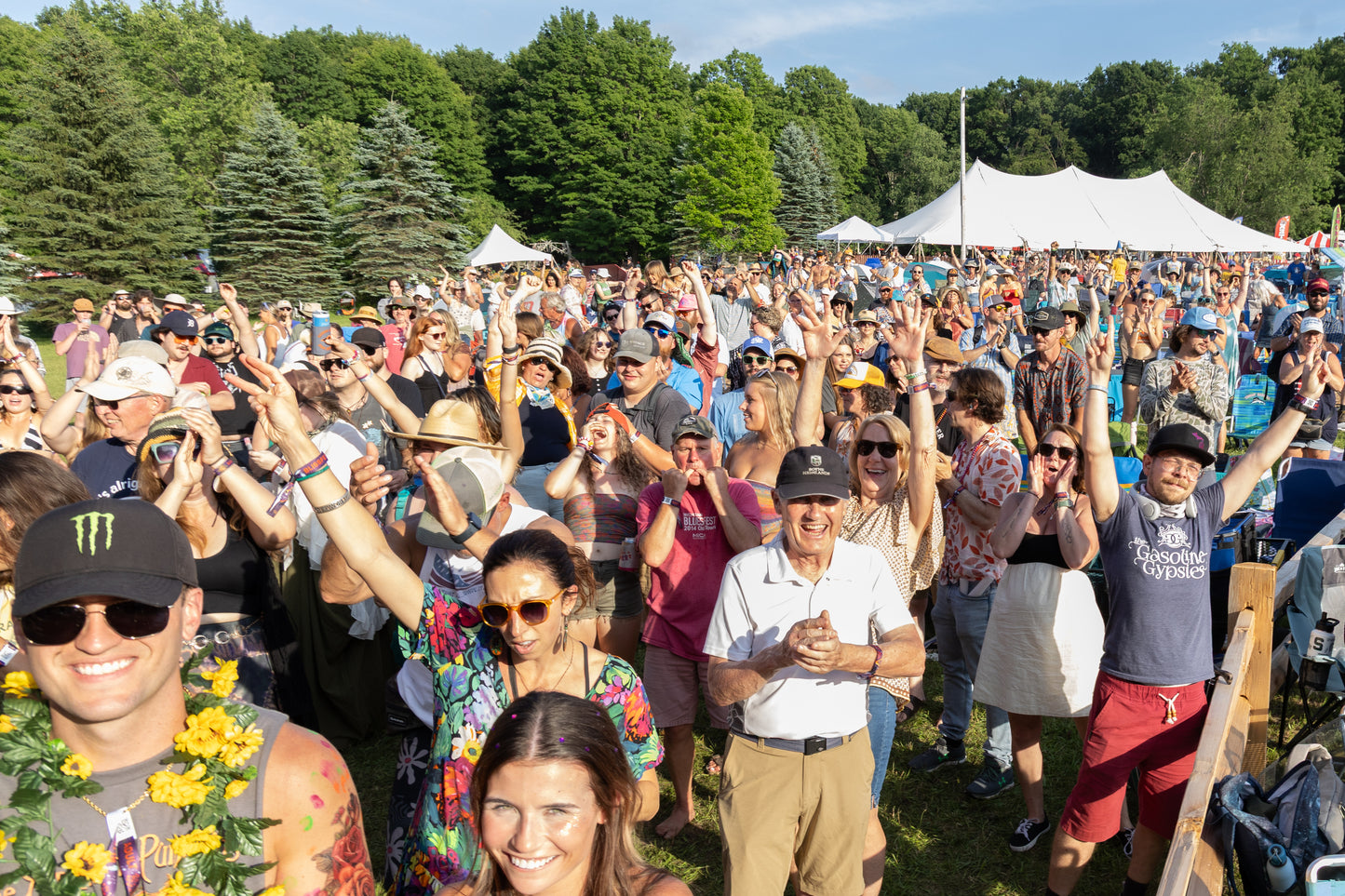 Large crowd of people at an outdoor event with trees and a white tent in the background.