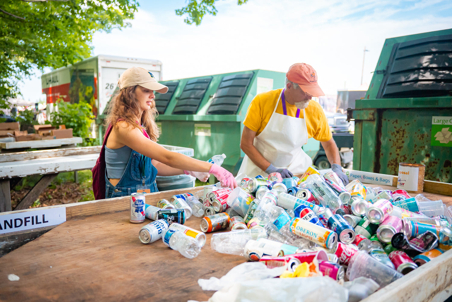 Two people sorting recyclables at a recycling station with bins and trees in the background.