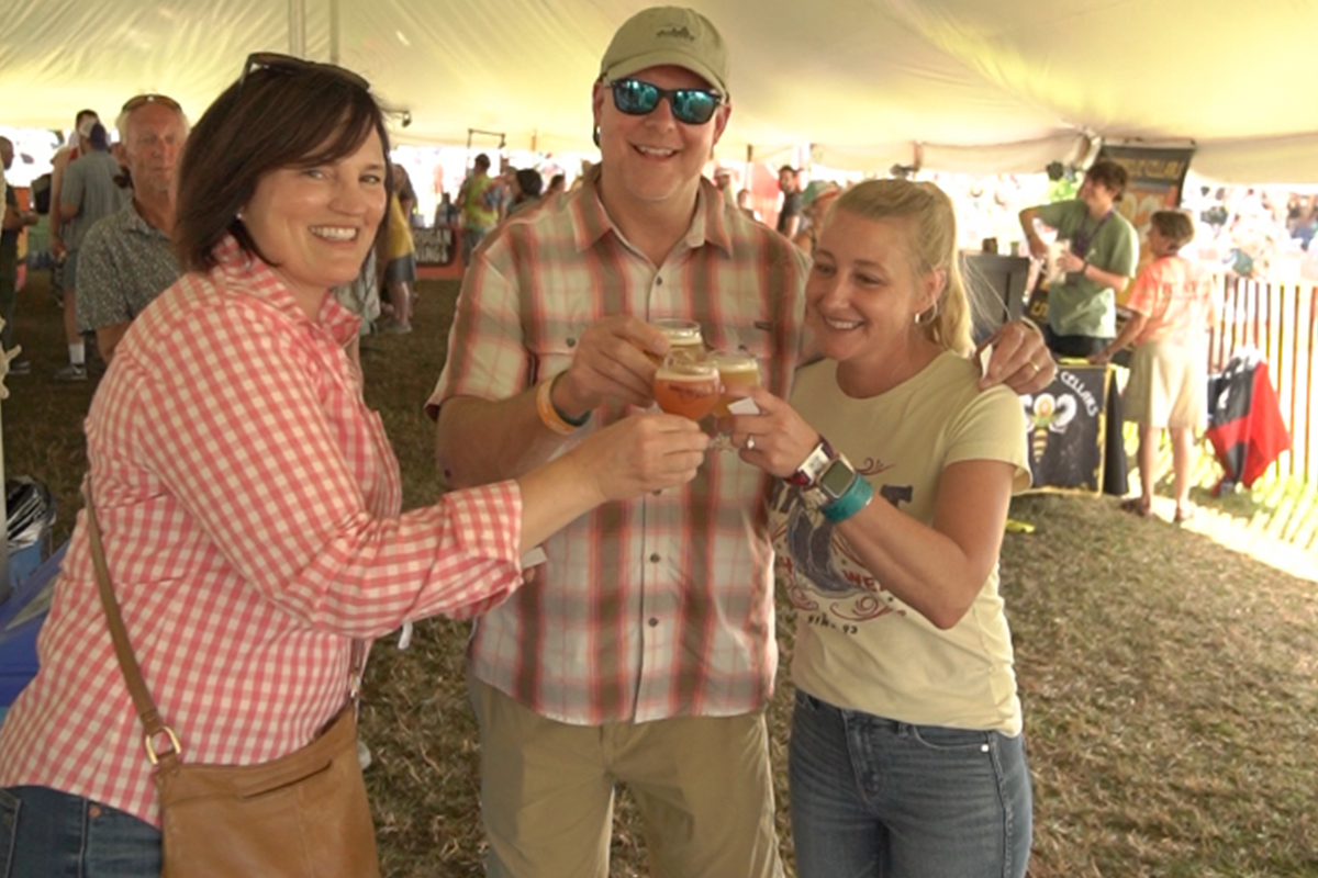 Three people toasting with drinks inside a tented area with other people in the background.
